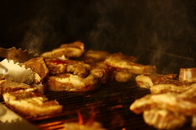 Several pieces of meat are being grilled on a barbecue, with smoke rising and a metallic tong in view, suggesting cooking in progress.