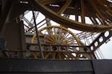 A close-up view of a complex industrial structure featuring large, yellow metallic wheels and beams. The geometric patterns of the metal framework create an intricate, mechanical aesthetic.