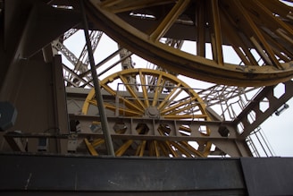 A close-up view of a complex industrial structure featuring large, yellow metallic wheels and beams. The geometric patterns of the metal framework create an intricate, mechanical aesthetic.