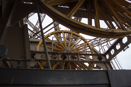 A close-up view of a complex industrial structure featuring large, yellow metallic wheels and beams. The geometric patterns of the metal framework create an intricate, mechanical aesthetic.