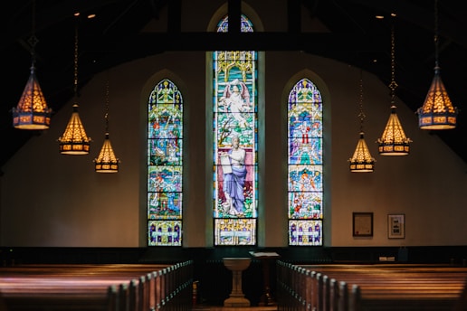 A dimly lit church interior focuses on three large stained glass windows that depict religious scenes. Ornate chandeliers hang from the ceiling, casting warm light. Wooden pews are arranged symmetrically, leading to a central altar or pulpit.