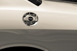 A close-up view of a car's fuel cap on the side of a light-colored vehicle. The metal cap is shiny and reflective, with the word 'FUEL' clearly marked on top.