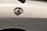 A close-up view of a car's fuel cap on the side of a light-colored vehicle. The metal cap is shiny and reflective, with the word 'FUEL' clearly marked on top.