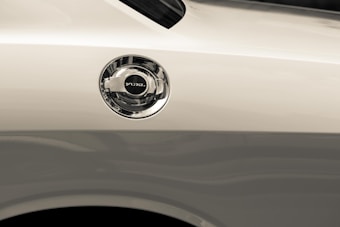 A close-up view of a car's fuel cap on the side of a light-colored vehicle. The metal cap is shiny and reflective, with the word 'FUEL' clearly marked on top.