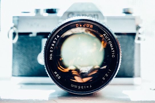 A close-up view of a vintage camera with the focus on the lens, showcasing details of the lens aperture and company's name. The camera body appears blurred in the background, highlighting the lens elements and text. Bright lighting illuminates the scene, giving a slightly overexposed, artistic effect.