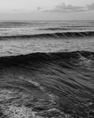 A black and white photograph of a vast ocean with waves rolling towards the shore. A lone surfer is visible in the distance, sitting on a board and waiting for a wave. Clouds are scattered across the sky, adding texture to the scene.