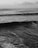 A black and white photograph of a vast ocean with waves rolling towards the shore. A lone surfer is visible in the distance, sitting on a board and waiting for a wave. Clouds are scattered across the sky, adding texture to the scene.