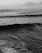 A black and white photograph of a vast ocean with waves rolling towards the shore. A lone surfer is visible in the distance, sitting on a board and waiting for a wave. Clouds are scattered across the sky, adding texture to the scene.