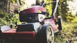 A red lawn mower sits on a grassy area surrounded by lush greenery. The focus is on the wheels and cutting deck, covered with remnants of grass. The background is slightly blurred with hints of sunlight filtering through the trees.