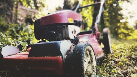 A red lawn mower sits on a grassy area surrounded by lush greenery. The focus is on the wheels and cutting deck, covered with remnants of grass. The background is slightly blurred with hints of sunlight filtering through the trees.
