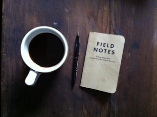 A creative workspace with a notebook, pen, and a cup of coffee on a wooden table.