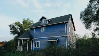 A two-story wooden house with blue siding stands amidst greenery. The house features white trim and a steep, dark shingled roof. A small gazebo with matching architectural details is located adjacent to the house. Trees with lush green foliage surround the property, and the sky above is mostly clear with soft lighting.