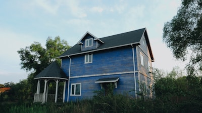 A two-story wooden house with blue siding stands amidst greenery. The house features white trim and a steep, dark shingled roof. A small gazebo with matching architectural details is located adjacent to the house. Trees with lush green foliage surround the property, and the sky above is mostly clear with soft lighting.