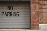A corrugated metal garage door with a 'No Parking' sign painted in black letters is adjacent to a brick wall. The bricks are varied in tone, displaying a red and brown color palette. The bottom edge of the door rests upon a concrete sidewalk.