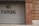 A corrugated metal garage door with a 'No Parking' sign painted in black letters is adjacent to a brick wall. The bricks are varied in tone, displaying a red and brown color palette. The bottom edge of the door rests upon a concrete sidewalk.