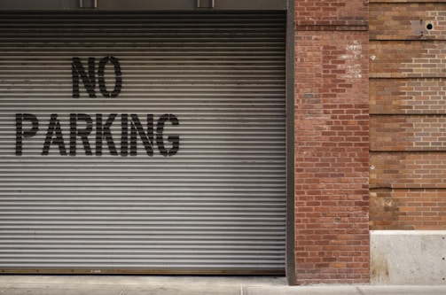 A corrugated metal garage door with a 'No Parking' sign painted in black letters is adjacent to a brick wall. The bricks are varied in tone, displaying a red and brown color palette. The bottom edge of the door rests upon a concrete sidewalk.