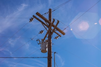 A wooden utility pole with crossbars stands tall against a clear blue sky. Several electrical insulators and wires are attached to the pole, connecting to a transformer. Lens flare creates hexagonal light reflections in the sky.