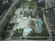An aerial view of an industrial area featuring several buildings, construction vehicles, machinery, and a variety of scattered equipment. The surrounding landscape includes trees and vegetation, with a main road traversing the site.