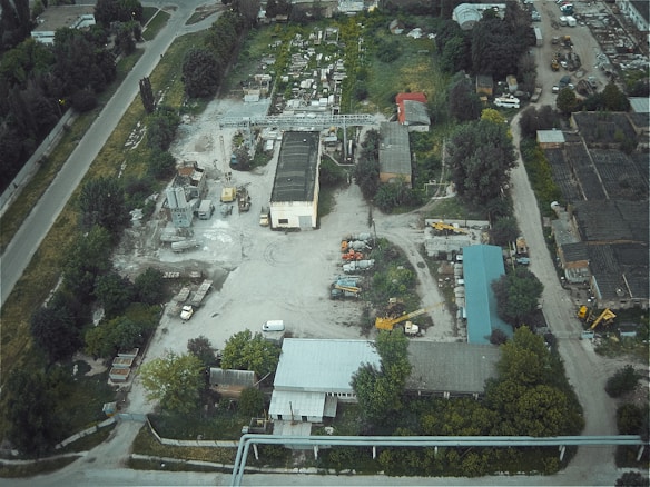 An aerial view of an industrial area featuring several buildings, construction vehicles, machinery, and a variety of scattered equipment. The surrounding landscape includes trees and vegetation, with a main road traversing the site.