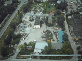 An aerial view of an industrial area featuring several buildings, construction vehicles, machinery, and a variety of scattered equipment. The surrounding landscape includes trees and vegetation, with a main road traversing the site.