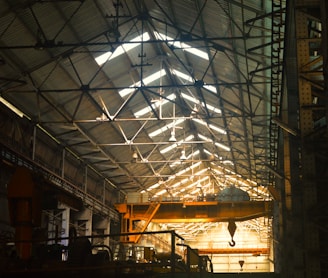 A large industrial warehouse with a high, metal, trussed ceiling. Sunlight filters through the windows, casting shadows across the interior. There is a prominent yellow overhead crane hanging from the ceiling, and various industrial equipment is visible throughout the space.