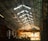 A large industrial warehouse with a high, metal, trussed ceiling. Sunlight filters through the windows, casting shadows across the interior. There is a prominent yellow overhead crane hanging from the ceiling, and various industrial equipment is visible throughout the space.