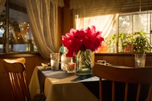 A cozy dining area bathed in warm sunlight, featuring a wooden table set with a dark tablecloth, a glass jug filled with vibrant red flowers as a centerpiece, and glasses filled with water. The background includes soft, billowing curtains and leafy green plants, enhancing the inviting atmosphere.