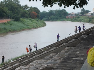 A riverbank features a gently flowing river beside lush greenery. Several people walk along a stepped embankment, engaged in various activities. Some stand near the water's edge, while others walk up the steps. Trees and buildings are visible in the background.