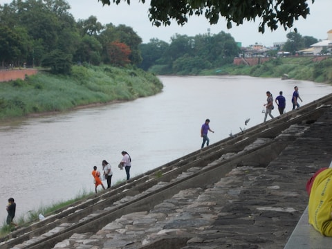 A riverbank features a gently flowing river beside lush greenery. Several people walk along a stepped embankment, engaged in various activities. Some stand near the water's edge, while others walk up the steps. Trees and buildings are visible in the background.