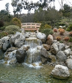 A peaceful garden scene features a small waterfall cascading over large rocks into a clear pond. A wooden footbridge arches over the waterfall, surrounded by lush greenery and trees in the background. The landscape combines natural elements in a harmonious setting.