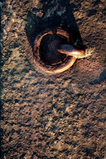 A textured stone surface with a rusted metal ring embedded in it. The metal ring is large and appears weathered, suggesting age and exposure to the elements. Sunlight casts a shadow, enhancing the texture and colors of both the metal and the stone.