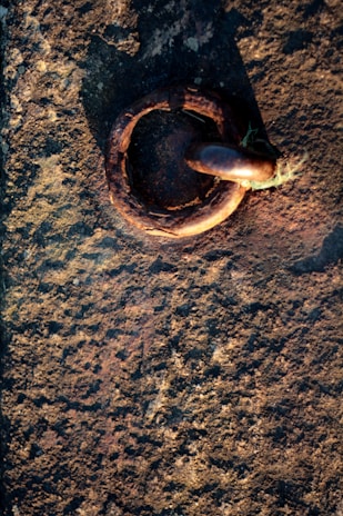 A textured stone surface with a rusted metal ring embedded in it. The metal ring is large and appears weathered, suggesting age and exposure to the elements. Sunlight casts a shadow, enhancing the texture and colors of both the metal and the stone.