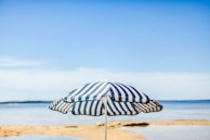 A beach umbrella with bold stripes planted firmly in the sand, waves gently rolling in the background.