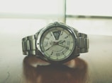 A sleek silver wristwatch rests on a wooden surface. The watch face displays the time with large numerals and features a day-date window. The metal bracelet complements the overall modern and sophisticated design, while the background appears blurred, emphasizing the watch.