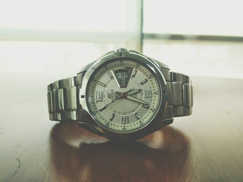 A sleek silver wristwatch rests on a wooden surface. The watch face displays the time with large numerals and features a day-date window. The metal bracelet complements the overall modern and sophisticated design, while the background appears blurred, emphasizing the watch.