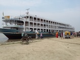 A large passenger ferry is docked by a sandy shore with several people boarding and disembarking. The ferry is multi-leveled and has windows along its sides. Nearby, there are people walking and a rickshaw positioned on the sandy area.