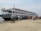 A large passenger ferry is docked by a sandy shore with several people boarding and disembarking. The ferry is multi-leveled and has windows along its sides. Nearby, there are people walking and a rickshaw positioned on the sandy area.