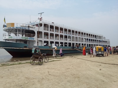 A large passenger ferry is docked by a sandy shore with several people boarding and disembarking. The ferry is multi-leveled and has windows along its sides. Nearby, there are people walking and a rickshaw positioned on the sandy area.