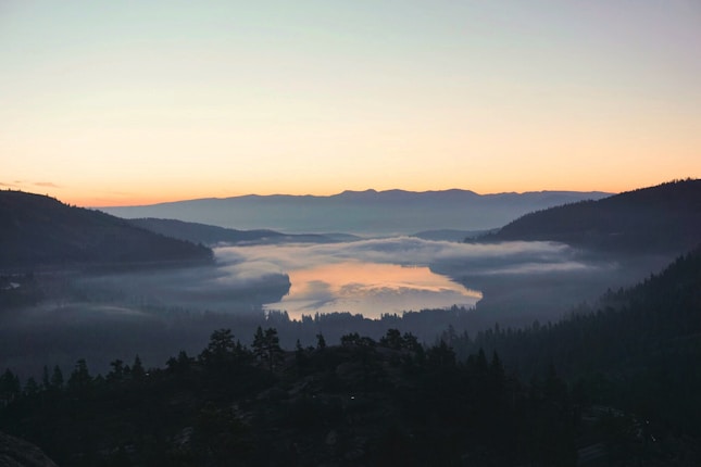 A serene mountain landscape at sunrise with a mist-covered lake in the center, surrounded by dense forests and hills. The sky transitions from a deep blue at the top to a warm orange near the horizon, creating a tranquil atmosphere.