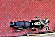 A top-down view of a black motorcycle parked on a tile surface with a red and white geometric pattern. The tiles have a distressed look, adding a vintage feel. The motorcycle features a large seat, metallic handlebars, and visible gauges on the dashboard.