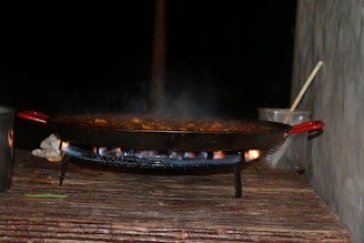 A large pan with red handles is situated on a stand over an open flame, with steam rising from the food being cooked inside. The setting appears to be outdoors, with a wooden surface beneath the pan and two metal or glass containers beside it, one containing a wooden spoon.