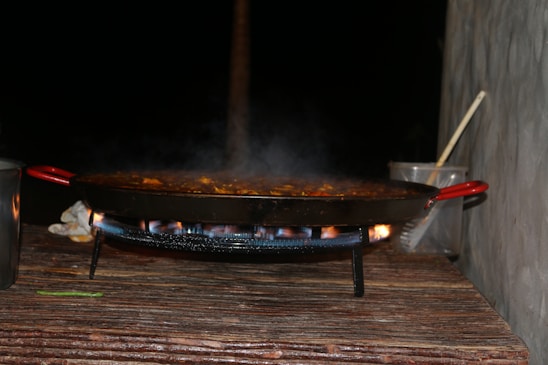 A large pan with red handles is situated on a stand over an open flame, with steam rising from the food being cooked inside. The setting appears to be outdoors, with a wooden surface beneath the pan and two metal or glass containers beside it, one containing a wooden spoon.