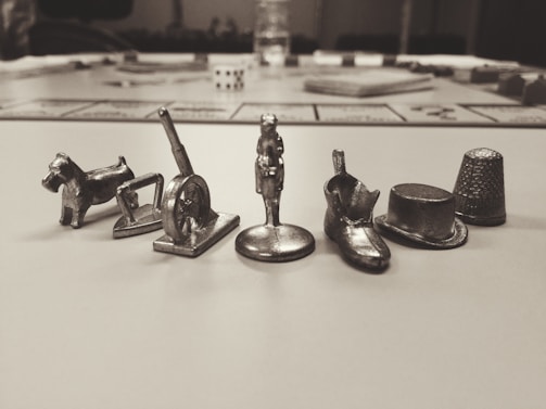Several small metallic game tokens representing different objects, such as a dog, a wheelbarrow, a cannon, a shoe, a hat, and a thimble, are lined up in focus on a blurred background that resembles a board game setup.