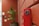 A close-up of a fire alarm or emergency device mounted on a red brick wall. The device is predominantly red with a metallic keyhole at the top. In the background, there is a blurred image of a green potted plant and a hint of a white doorway.