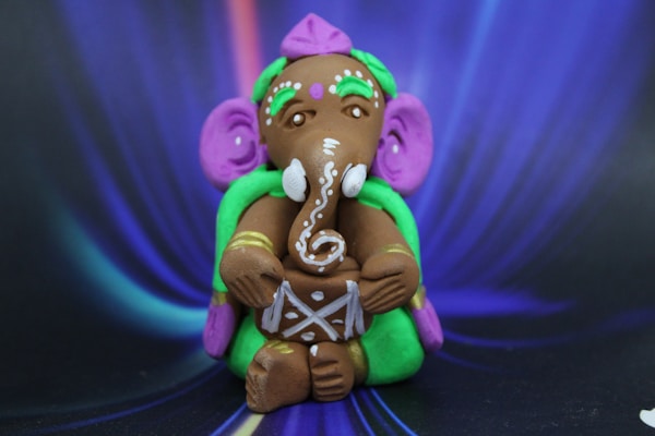 A colorful figurine of Lord Ganesha made of clay, featuring intricate patterns and vibrant colors. The deity is adorned with a detailed headdress and holds a musical drum. The background has a swirling pattern in shades of blue and purple.