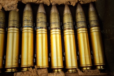 A collection of brass-colored artillery shells arranged neatly in a crate. The shells are cylindrical with pointed tips and detailed markings inscribed on them. They appear well-maintained and are placed closely together in a row, surrounded by padding material.