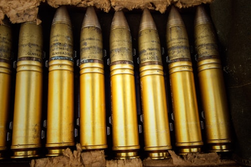 A collection of brass-colored artillery shells arranged neatly in a crate. The shells are cylindrical with pointed tips and detailed markings inscribed on them. They appear well-maintained and are placed closely together in a row, surrounded by padding material.
