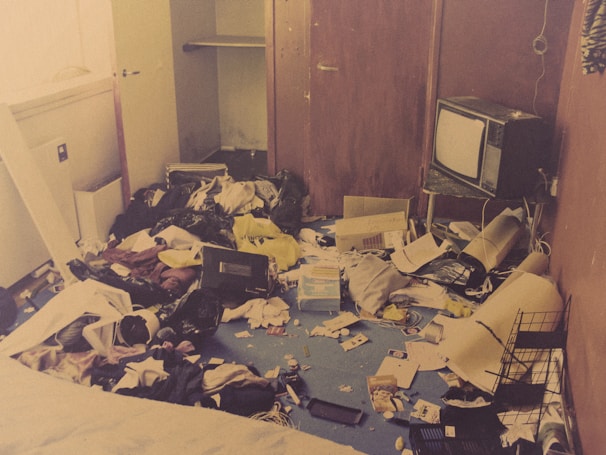 A cluttered room with clothes, papers, and miscellaneous items scattered across a blue carpet. A vintage television sits atop a small stand against a wood-paneled wall. An open closet door reveals an empty hanging rod. Various boxes, cables, and debris are strewn around, contributing to the disorganized appearance.