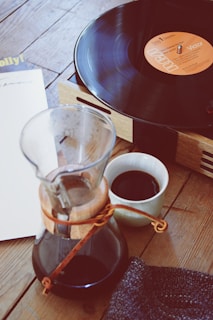 A wooden tabletop holds a record player with a vinyl playing. Nearby, there's a glass carafe with coffee and a small cup filled with coffee. A piece of fabric lies beside the cup. The scene suggests a cozy, relaxed setting.