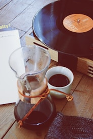 A wooden tabletop holds a record player with a vinyl playing. Nearby, there's a glass carafe with coffee and a small cup filled with coffee. A piece of fabric lies beside the cup. The scene suggests a cozy, relaxed setting.